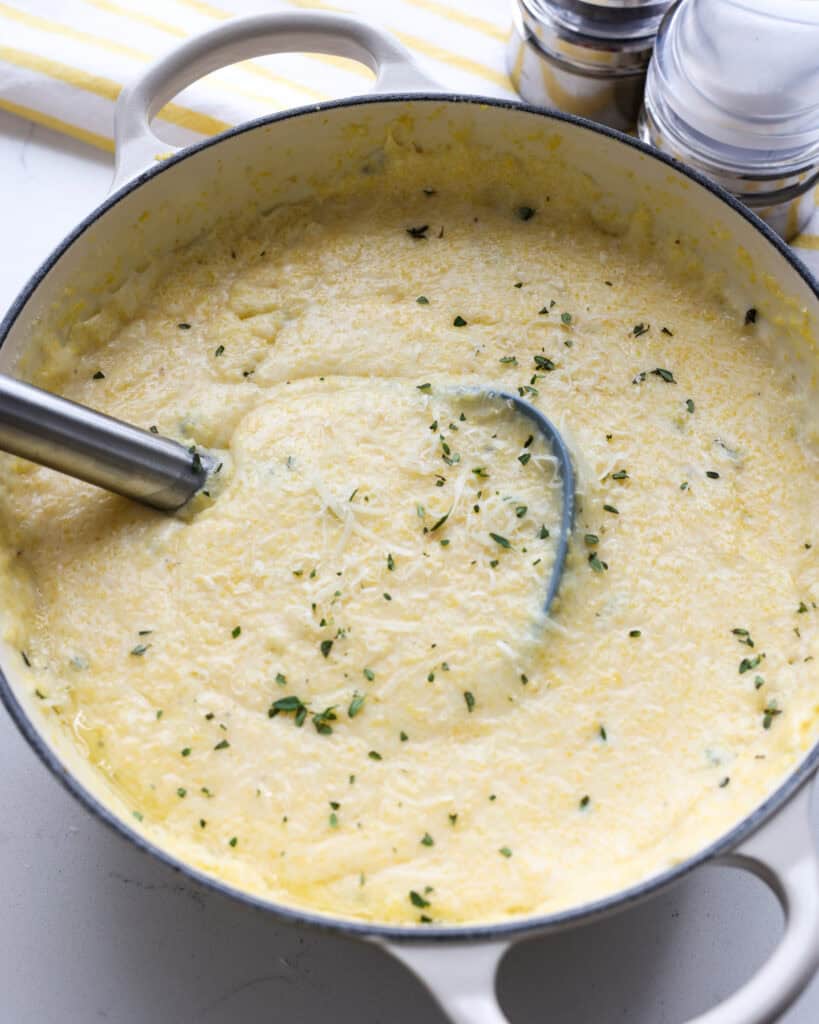 A white Dutch oven and ladle full of creamy polenta garnished with fresh thyme.