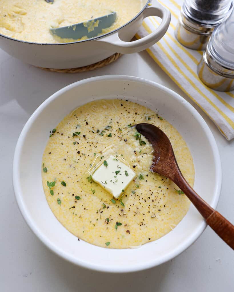 A single serving white bowl full of creamy polenta with a pat of butter in it. There is a salt and peppr shaker in the background and a yellow and white striped towel.