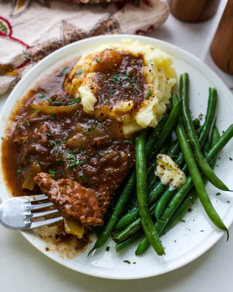 A slice of cube steak on a fork on a plate with the steak, mashed potatoes, gravy and French style green beans.