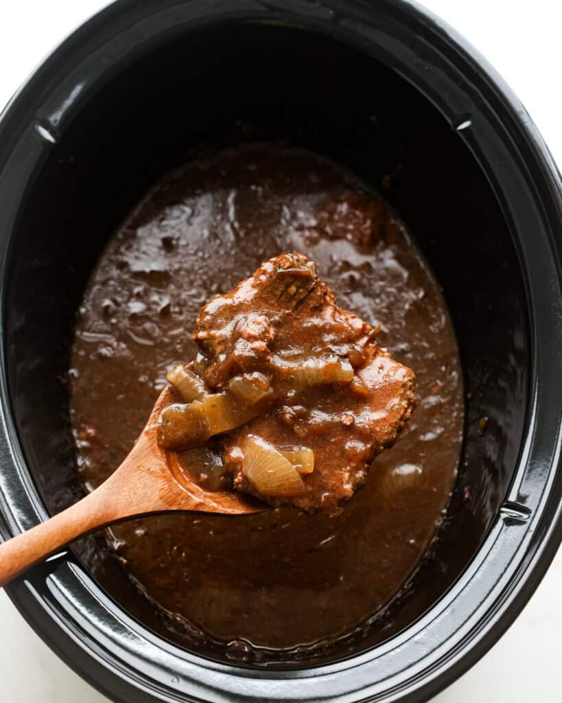A wooden spatula holding a cube steak over a crock pot full of cube steak and gravy.