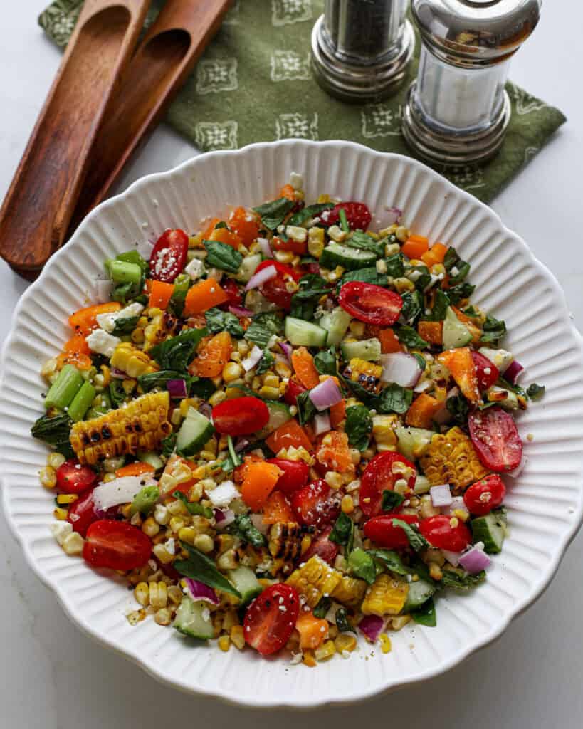 A bowl full of grilled corn salad with tongs and salt and pepper shaker  in the background