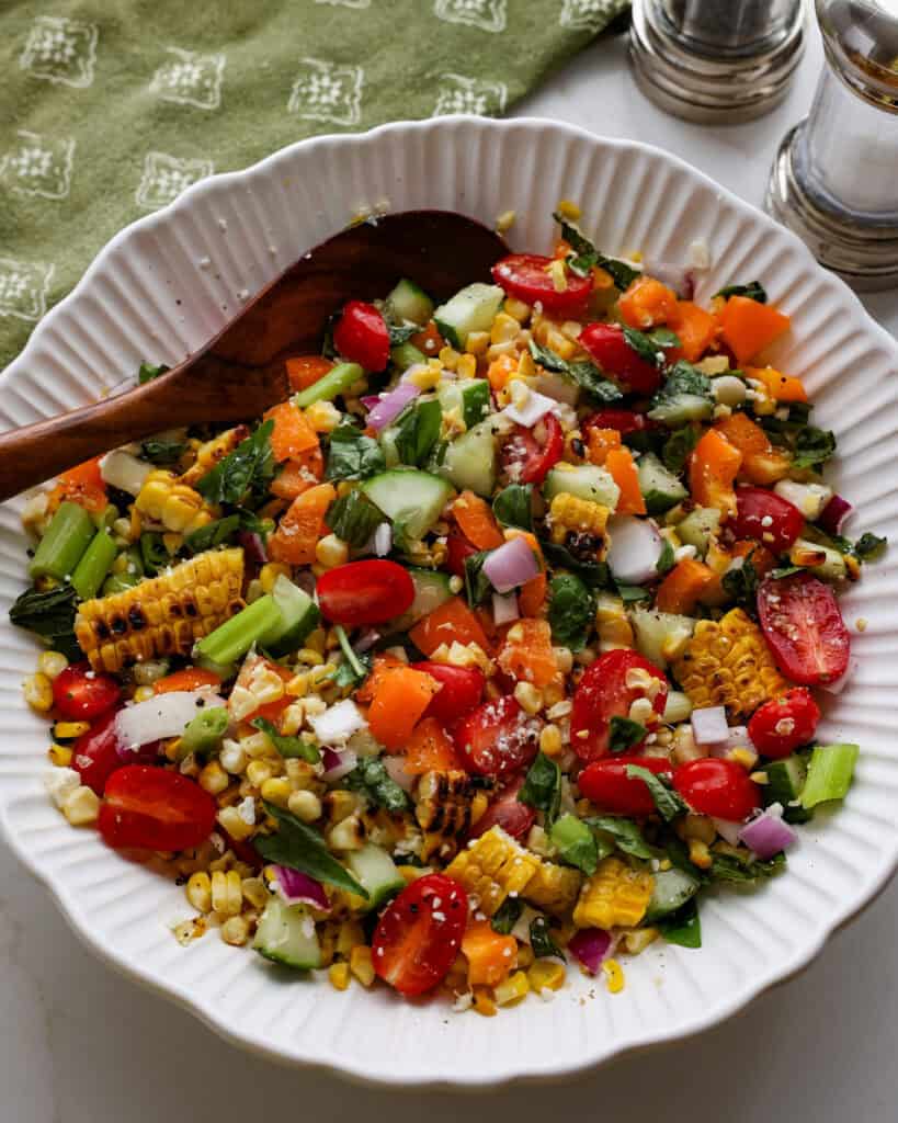 An overhead view of a white serving bowl full of grilled corn salad with a wooden serving spoon in it. 