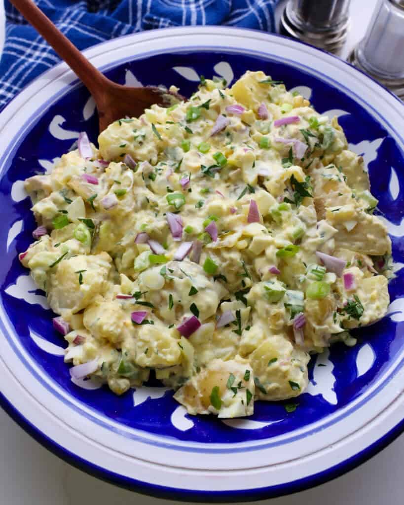 A large blue and white serving bowl and a wooden spoon full of mustard potato salad. 