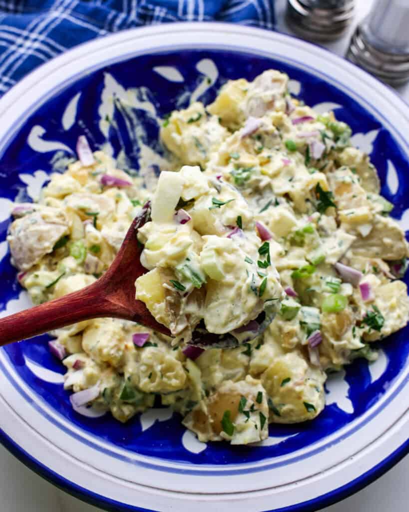 A heart spoon full of mustard potato salad over a large blue and white serving bowl full of mustard potato salad. 