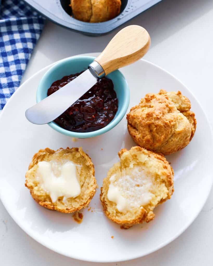 A plate with a split sour cream biscuit with melted butter on it. There is a small bowl of raspberry jam in the background. 