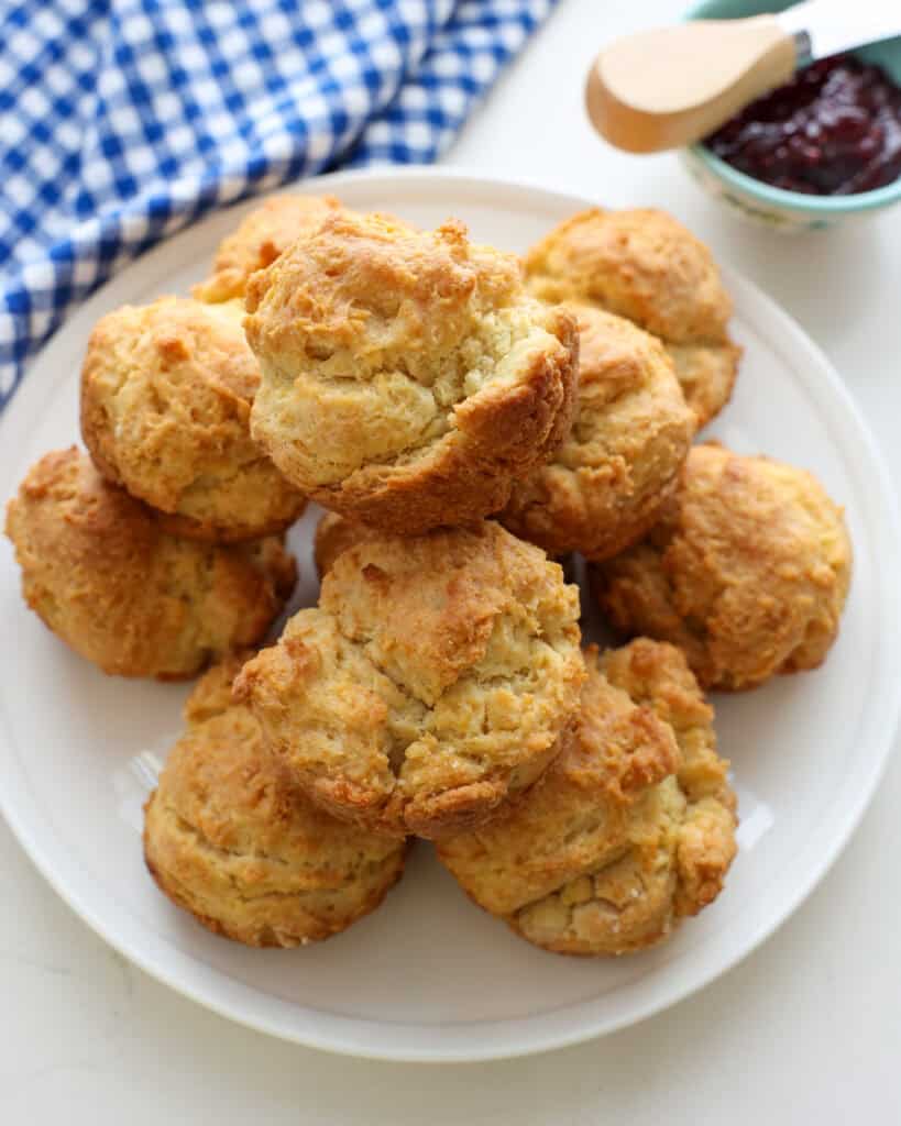 A serving plate full of sour cream biscuits with a kitchen towel and a bowl full of raspberry jam in the background. 
