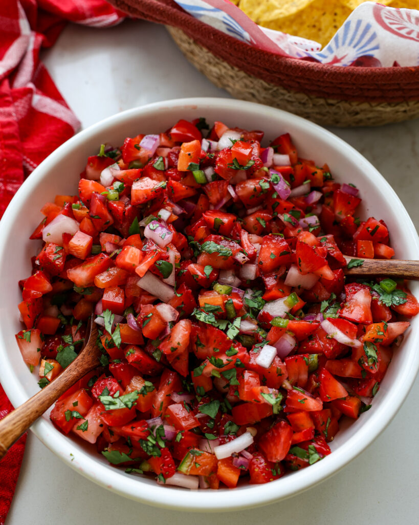 A bowl of salsa with two wooden spoons in it with chips in the background. 
