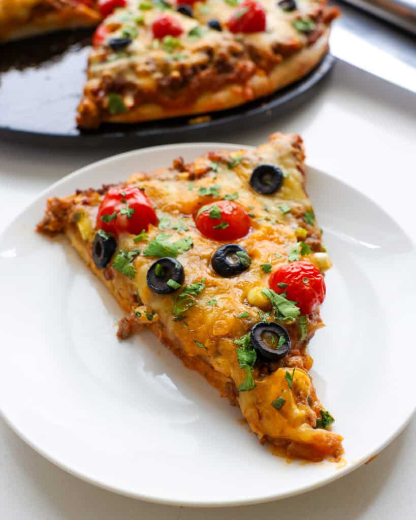 A slice of taco pizza on a single serving plate with the rest of the pizza in the background. 