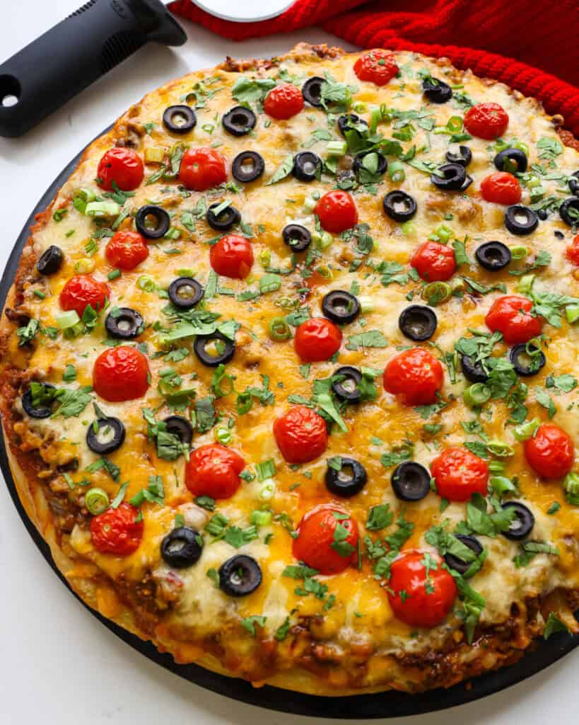 An overhead view of a taco pizza on a pizza stone with a pizza cutter and red towel in the background. 