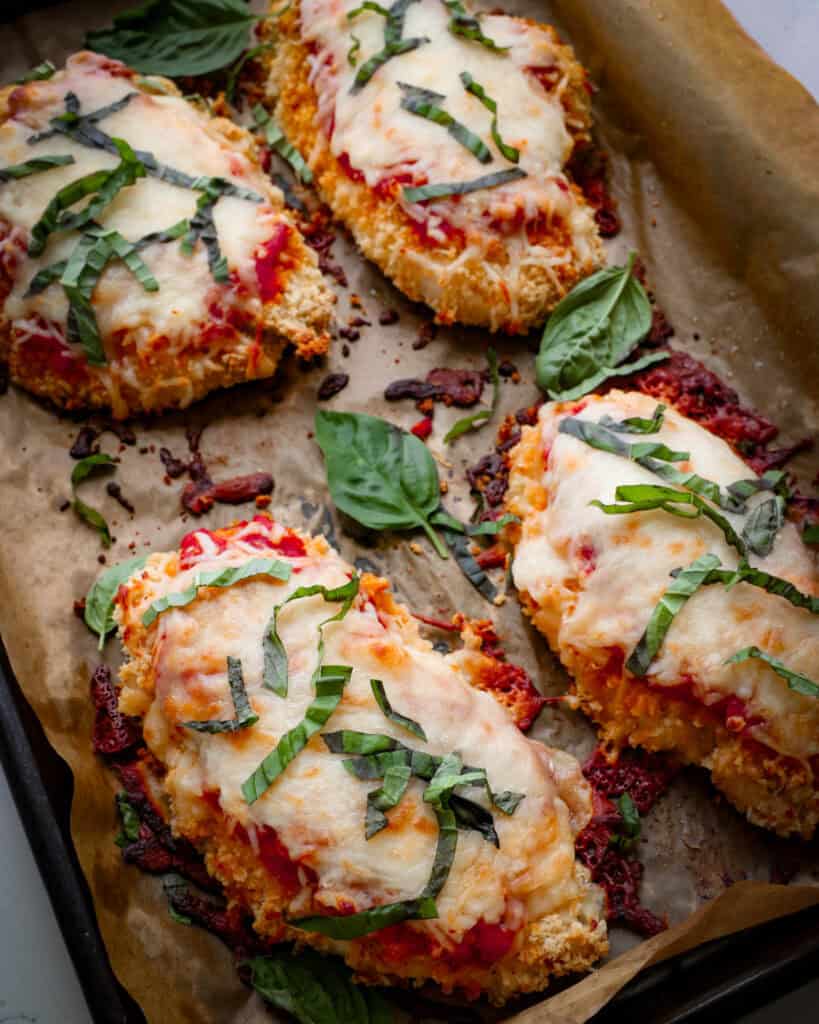 A close up view of baked chicken parmesan with basil ribbons on a parchment covered baking sheet. 