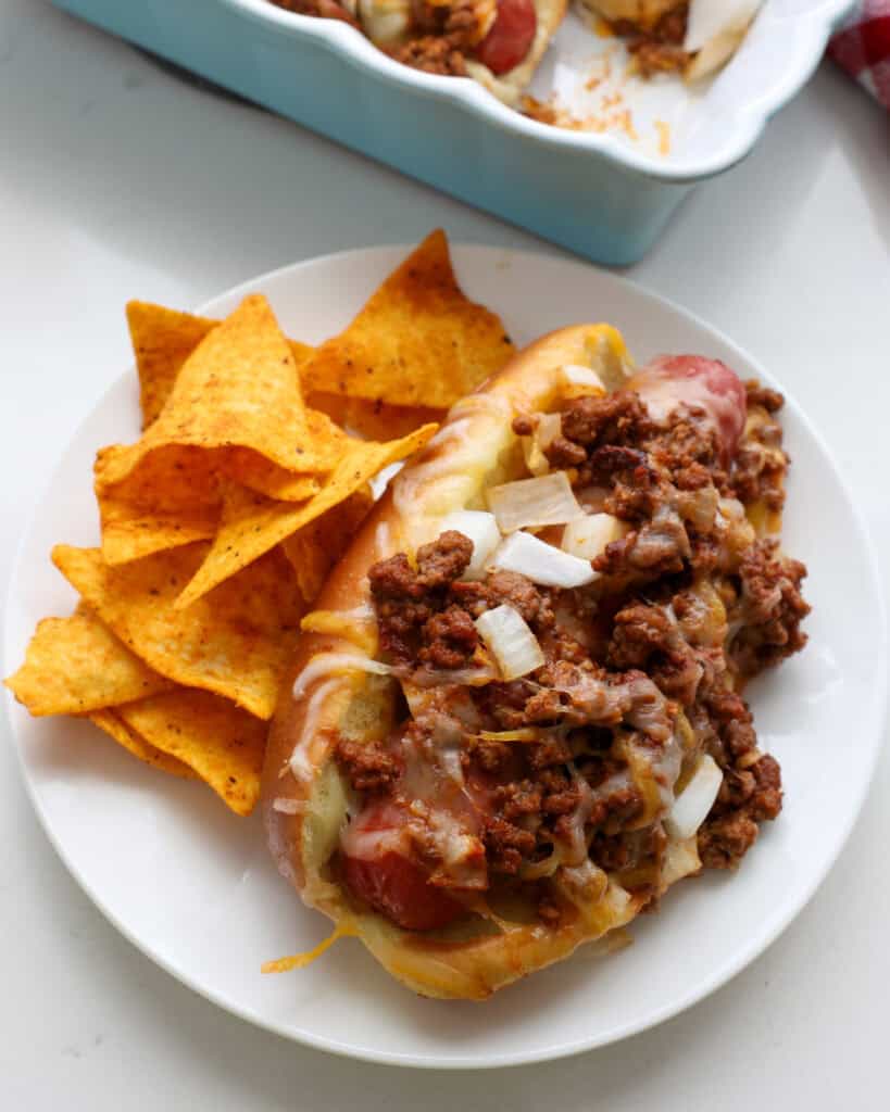 A chili dog with onions and cheese on a plate with spicy tortilla chips.