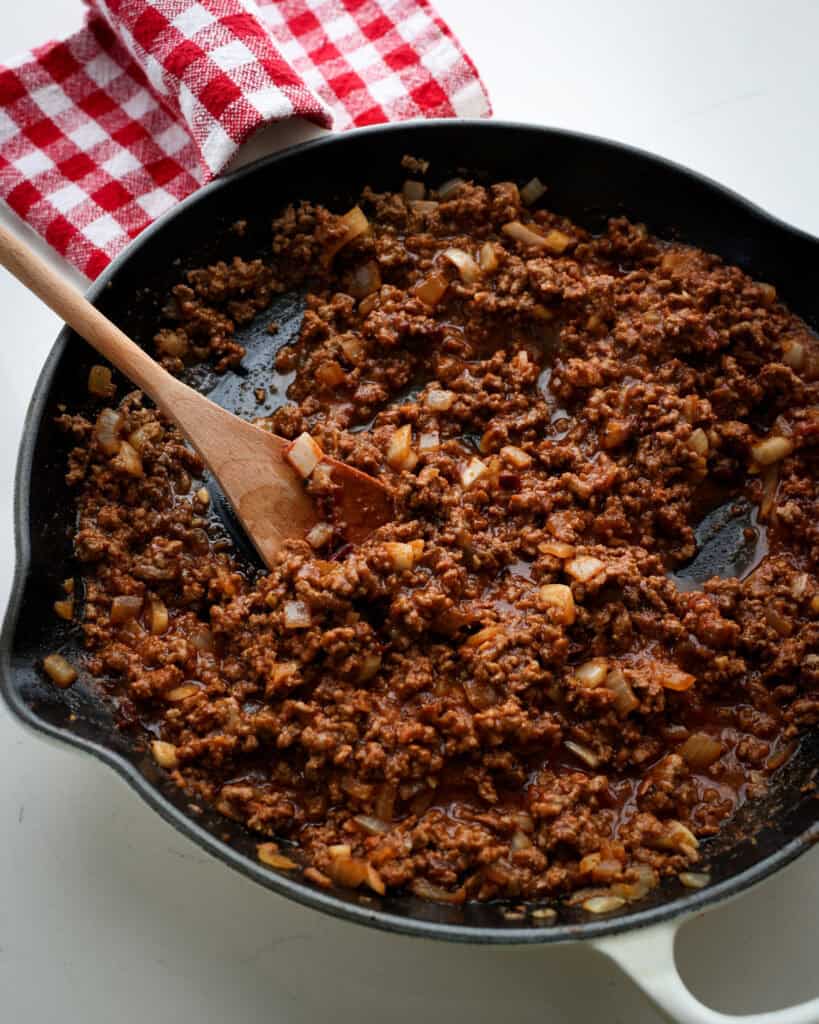A white cast iron skillet and a wooden spoon full of hot dog chili.