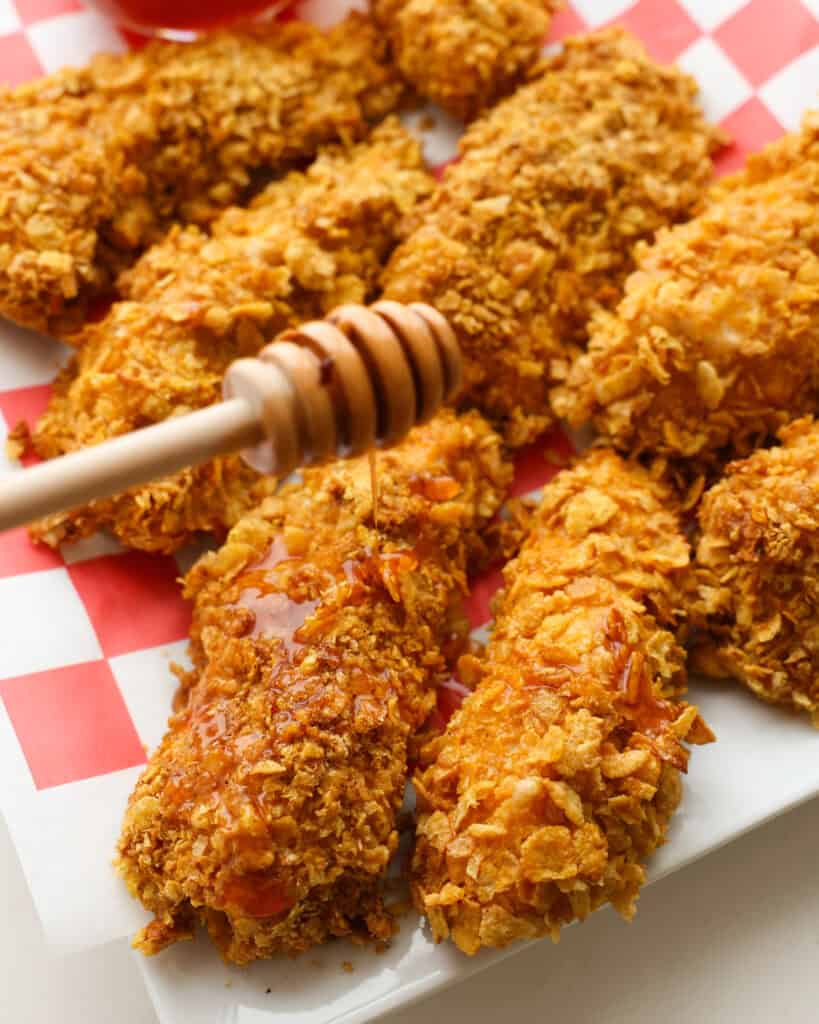 A platter full of cornflake breaded chicken tenders drizzled with hot honey sauce. 