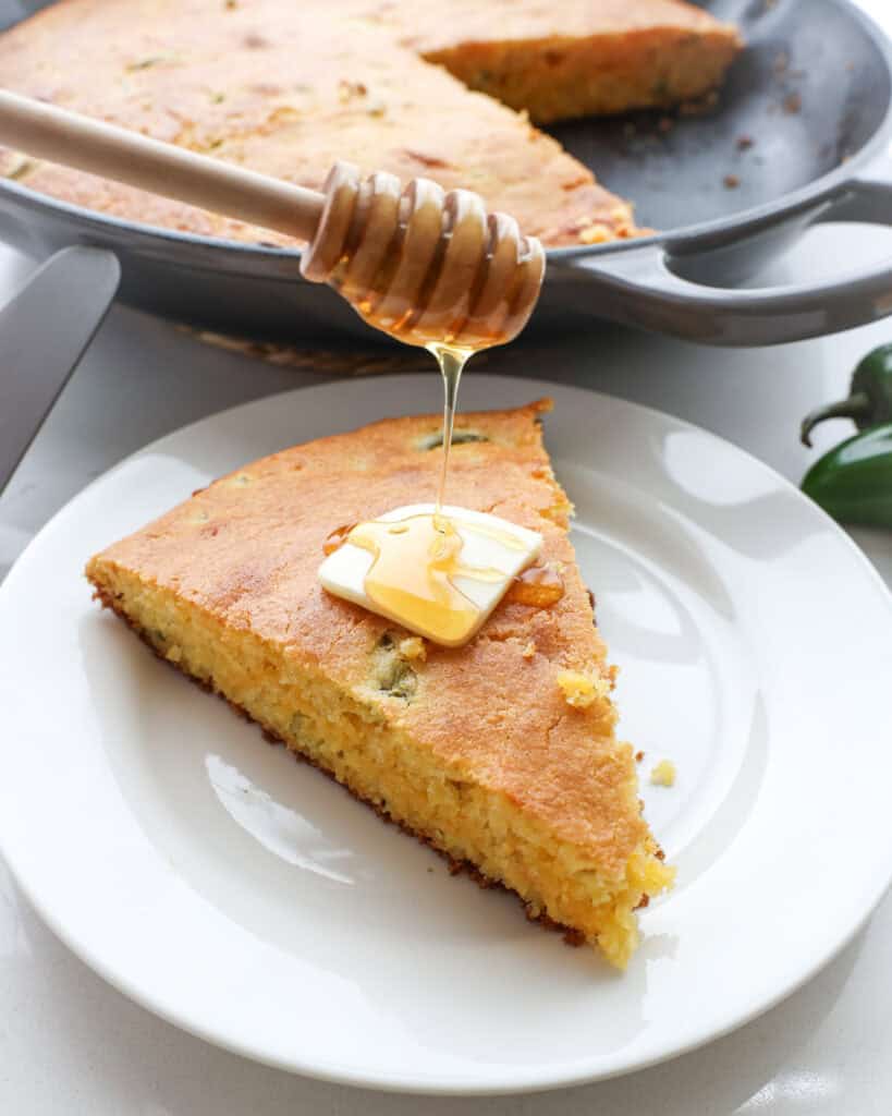 A slice of jalapeno cornbread on a single serving plate. A pat of butter and drizzled honey on the cornbread.