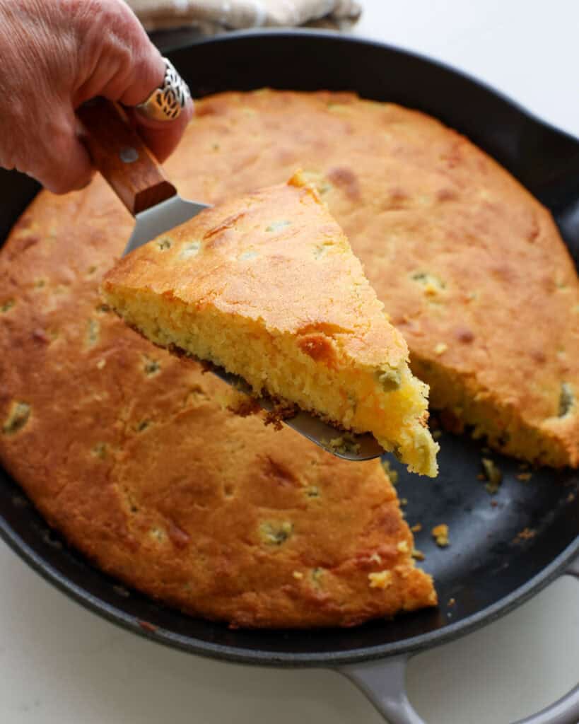 A slice of jalapeno cornbread on a serving spatula over a cast iron skillet of the jalapeno cornbread.