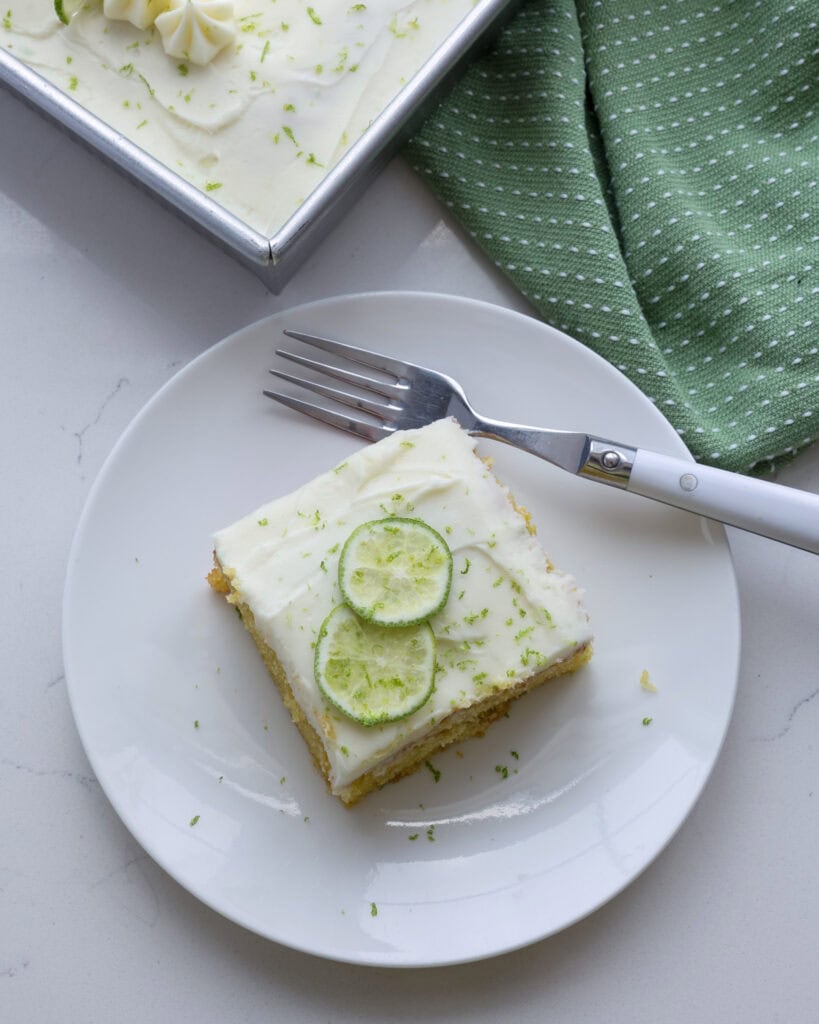 An overhead view of a piece of key lime cake on a serving plate with a fork and the rest of the cake in the background. 