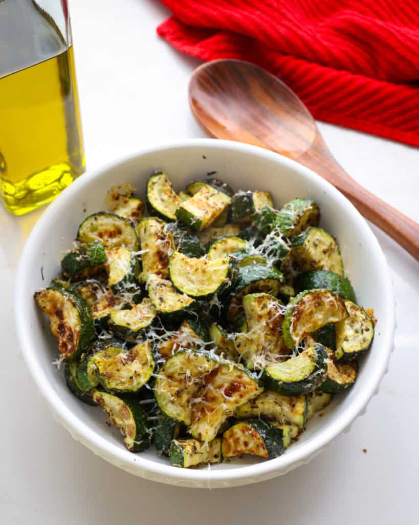 A serving bowl full of roasted zucchini with a wood spoon and olive oil in the background.
