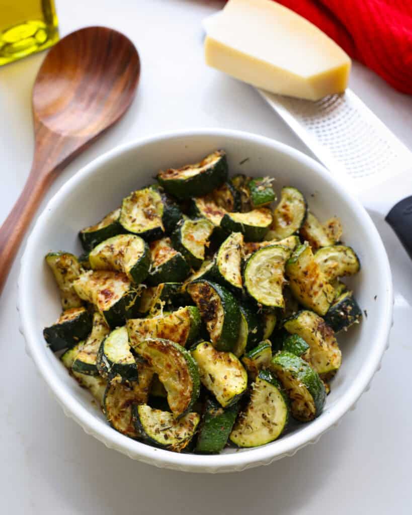 A white serving bowl full of roasted zucchini with a block of Parmesan cheese, a microplane grater, wooden spoon, and olive oil in the background.