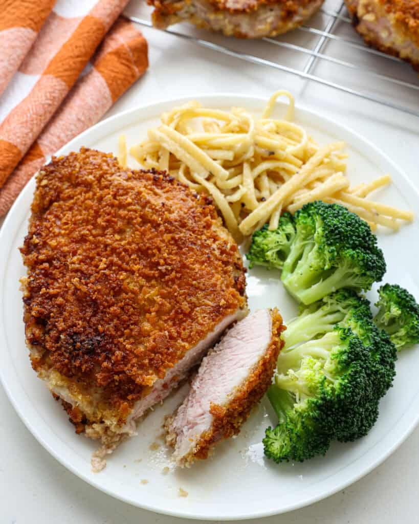 A Parmesan crusted pork chop with a slice cut off on a plate with steamed broccoli and Parmesan pasta. 
