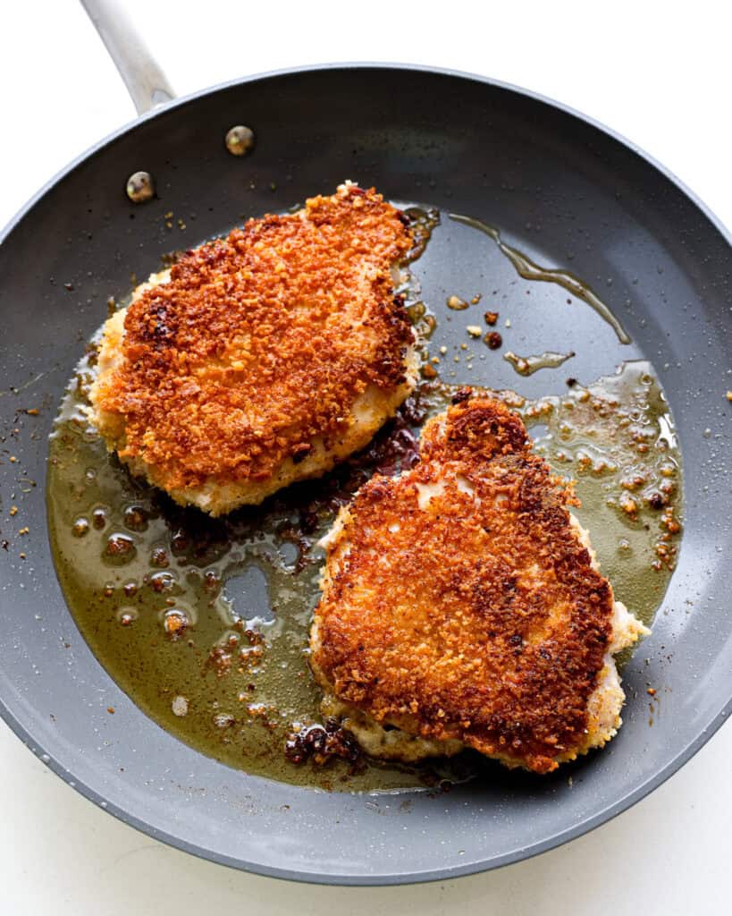 Two Parmesan crusted pork chops frying in a pan with oil. 