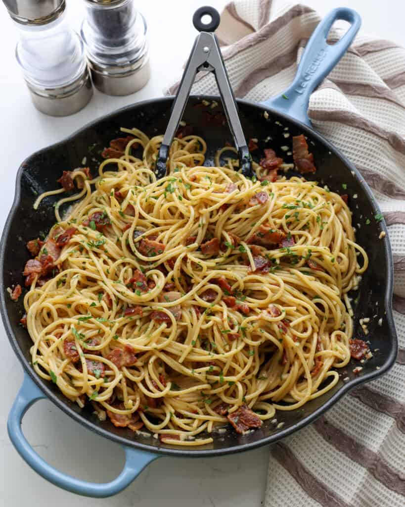 An overhead view of a blue skillet full of pasta carbonara with a salt and pepper shaker in the background.