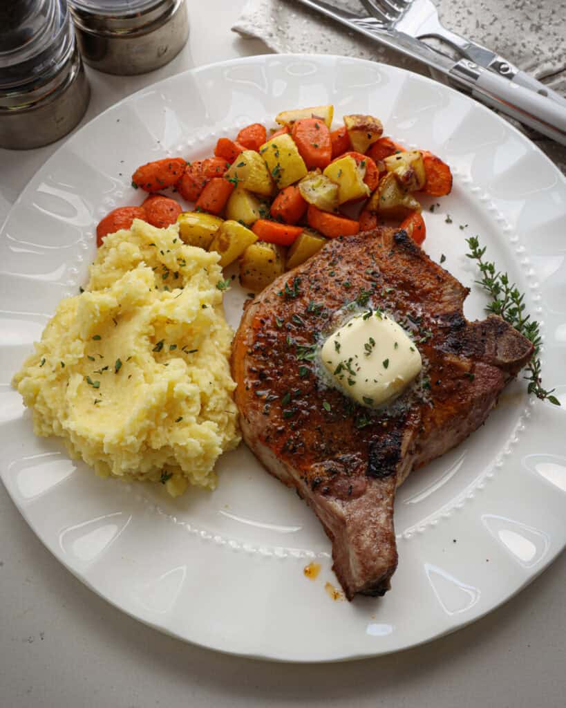 An oven baked pork chop on a white serving plate with mashed potatoes and yellow sqaush with carrots.