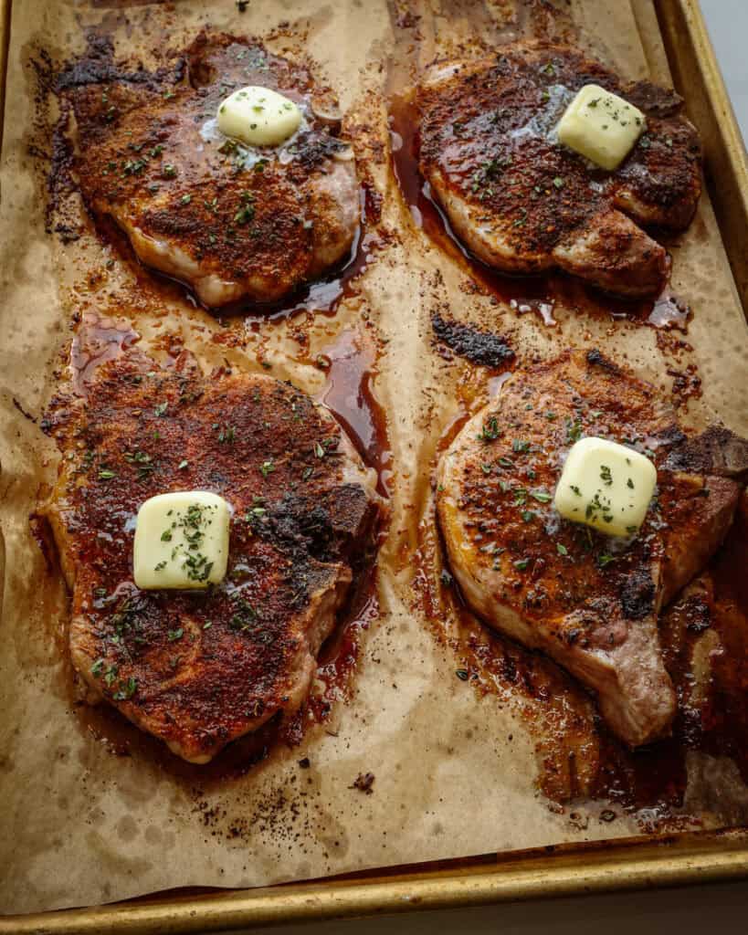 Four baked pork chops topped with a butter pat on a baking sheet covered with parchment paper.