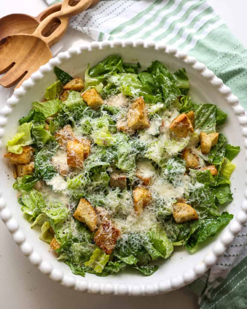 An overhead view of Caesar chicken salad in a white serving bowl with wooden serving tongs in the background.