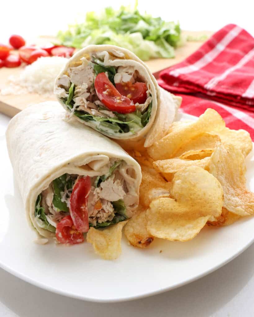 A chicken Caesar wrapped stacked on a white plate with chips on the side. The ingredients to make more chicken Caesar wraps in the background.