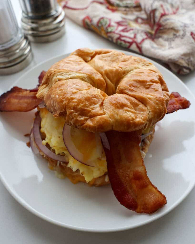 A close up view of a croissant breakfast sandwich on a single serving plate with a napkin and salt and pepper shaker in the background.