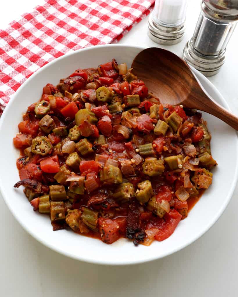 Okra and tomatoes in a white serving bowl with a wooden spoon. Salt and pepper shaker are in the background.