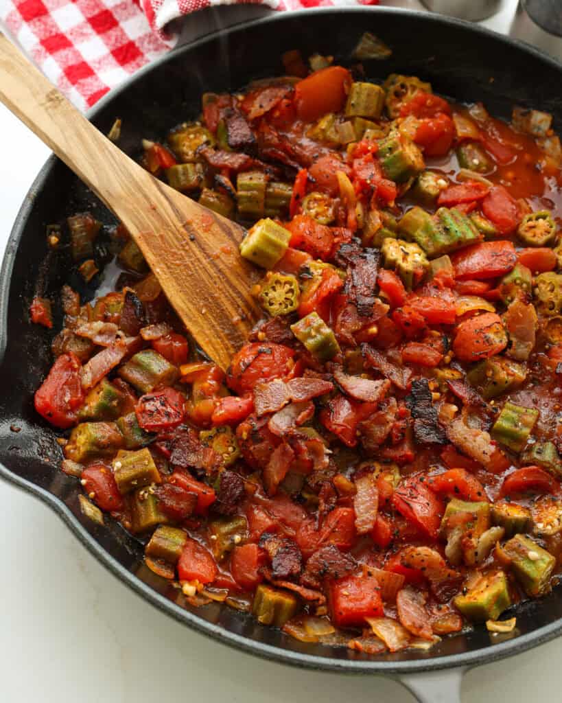 A close up view of okra and tomatoes in a skillet with a wooden spatula.