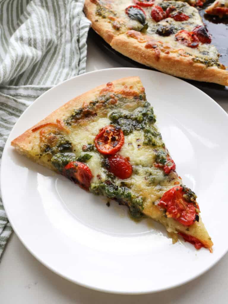 A slice of pesto pizza on a white single serving plate with the rest of the pizza in the background.