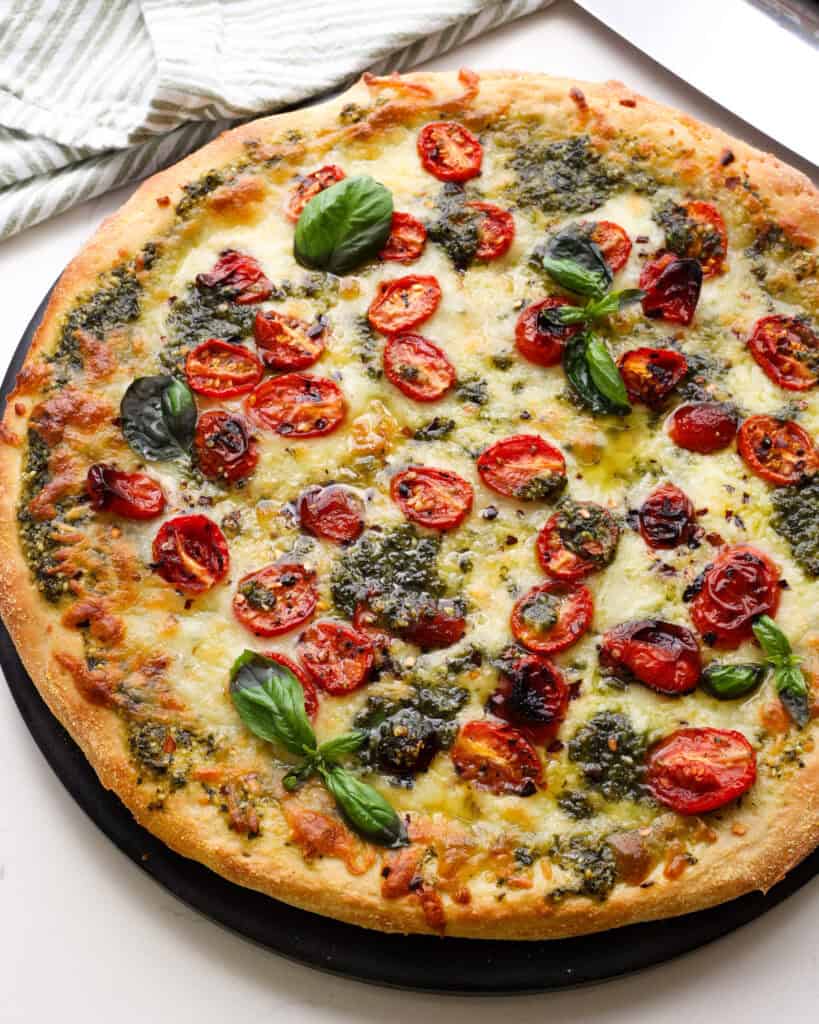 An uncut pesto pizza with roasted tomatoes fresh basil and mozzarella.