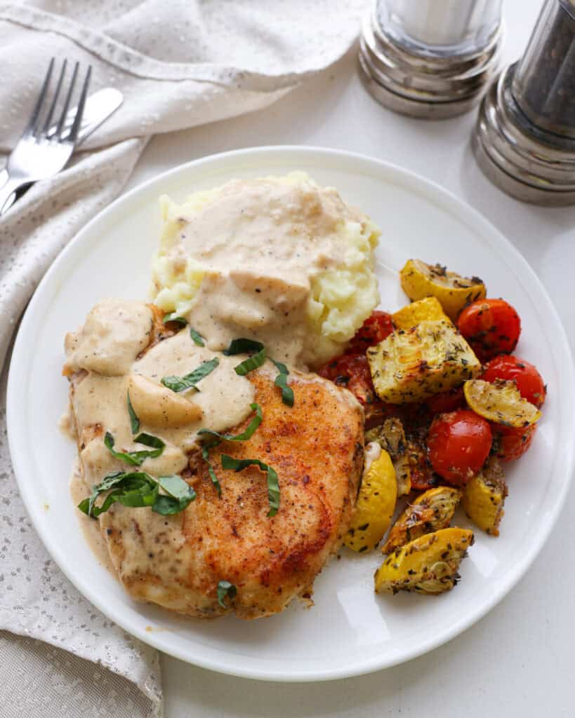 Creamy garlic chicken, mashed potatoes, and yellow squahs with tomatoes on a single white serving plate.