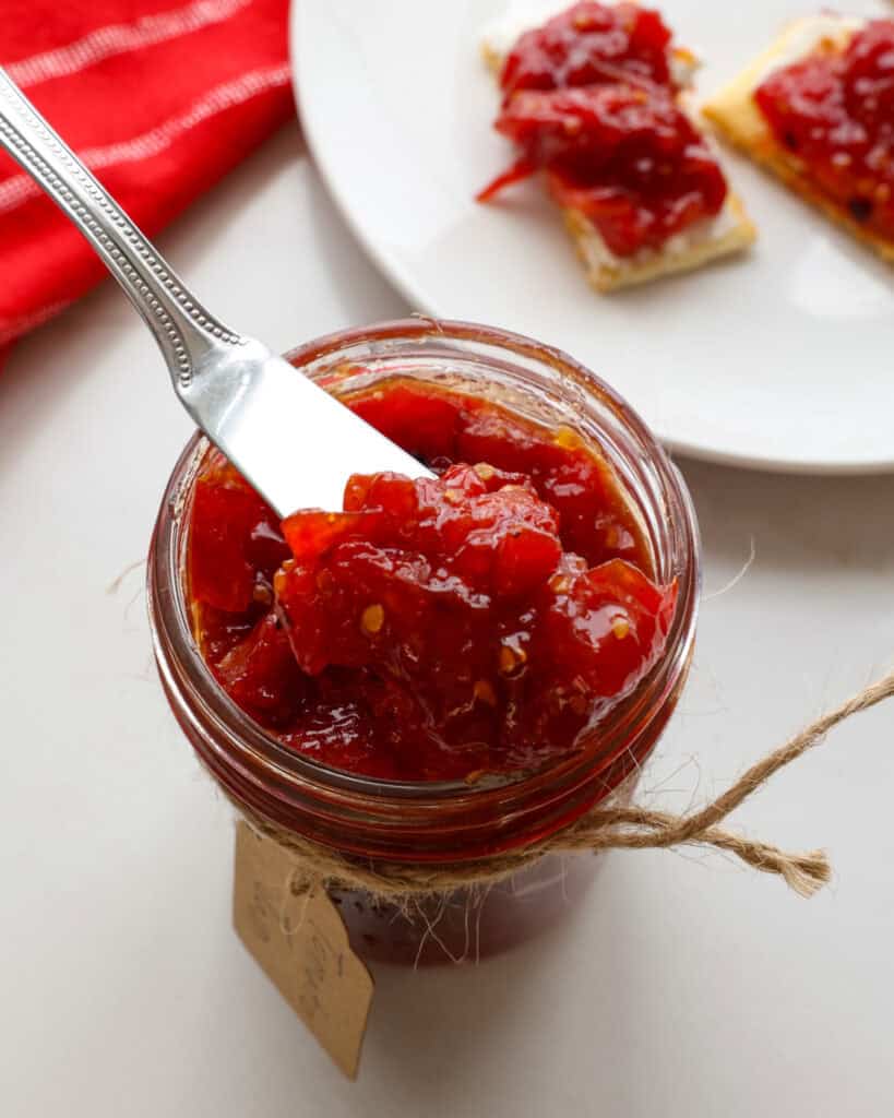 A close up view of tomato jam on a butter knife in a mason jar full of tomato jam.  