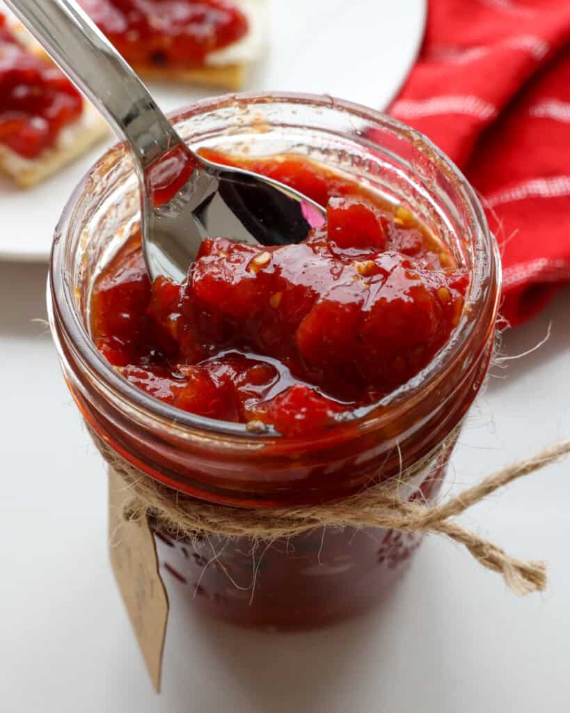 A spoon full of tomato jam in a mason jar full of tomato jam with a red kitchen towel and crackers with cream cheese and tomato jam in the background. 