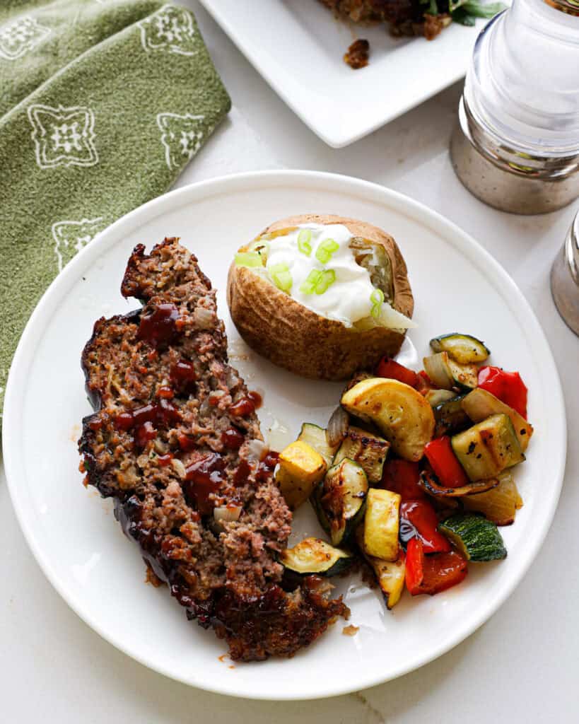 A large slice of air fryer meatloaf on a white serving plate with a baked potato with sour ceam and sauted zucchini, yellow squash and bell pepper. 