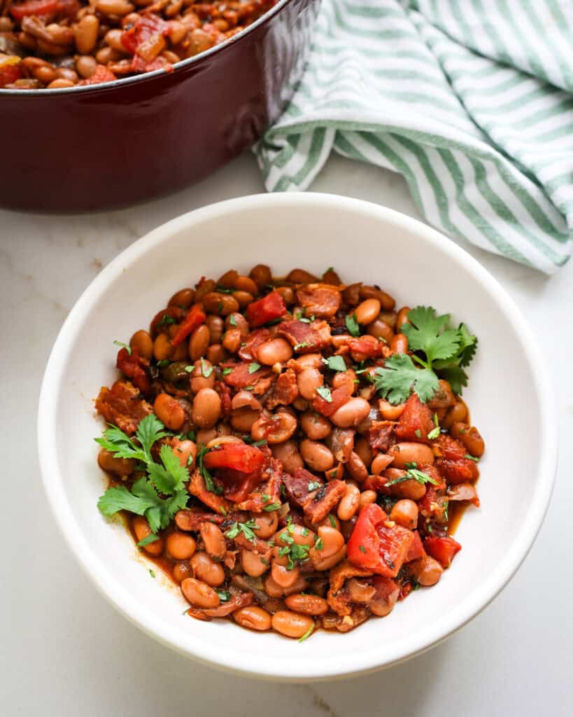 A white serving bowl full of borracho beans with a pot full of beans in the back ground.
