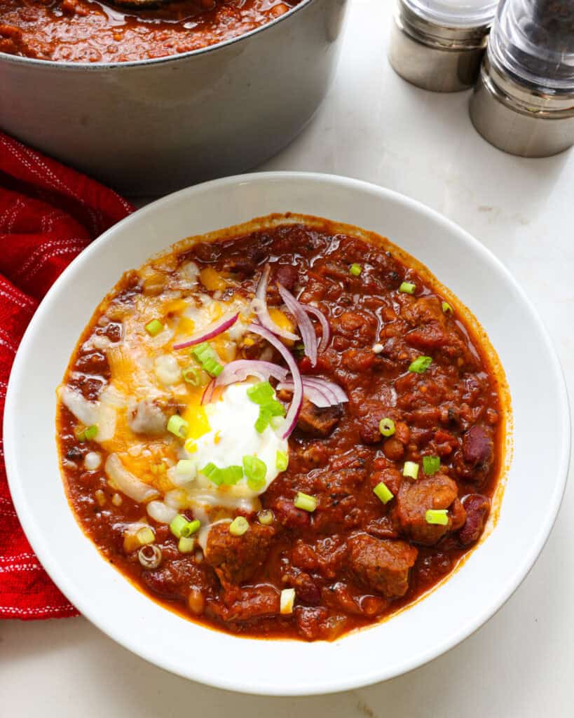 A white serving bowl full of brisket chili topped with sour cream, cheddar cheese, red onions, and green onions.