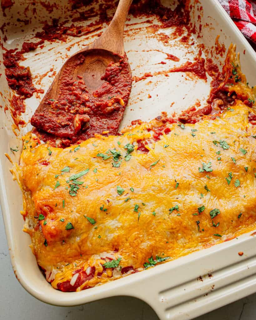 A casserole half full of enchiritos with a wooden spatula in it. 