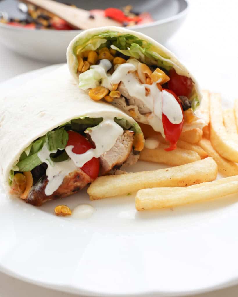 A half wrap stacked on top of another half wrap with French fries on the side and the skillet with the remainging corn, black beans and tomatoes in the background.