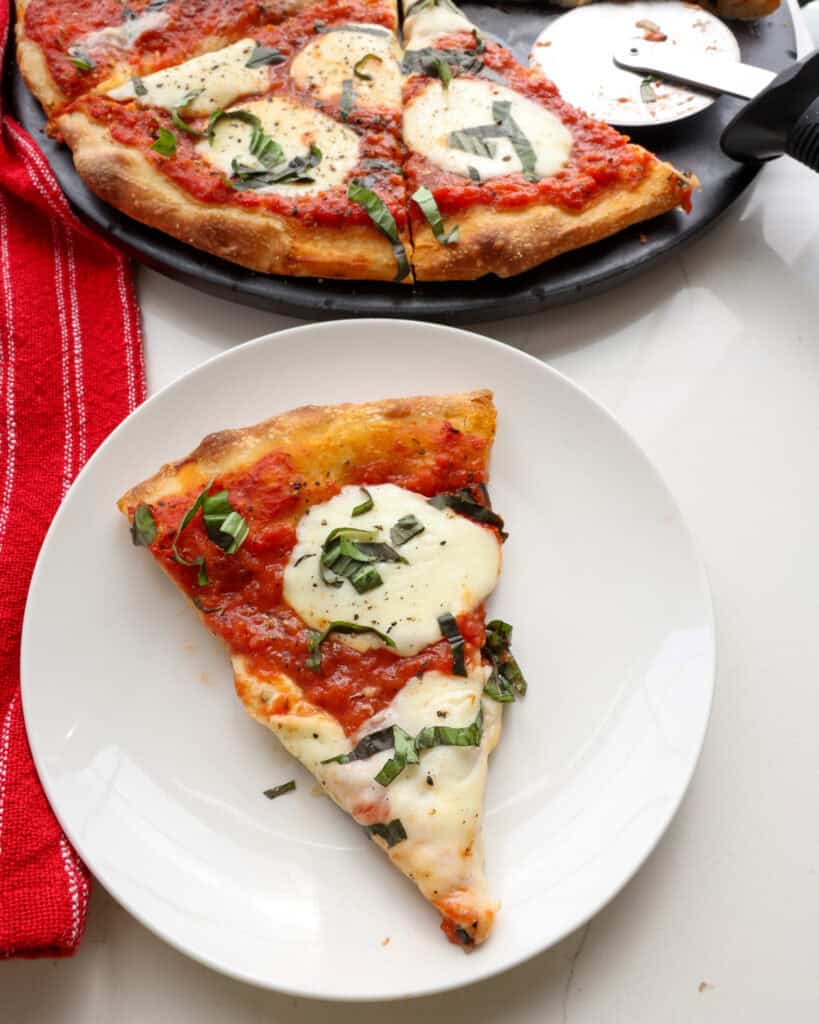 A slice of margherita pizza on a white serving plate with the rest of the pizza in the background.