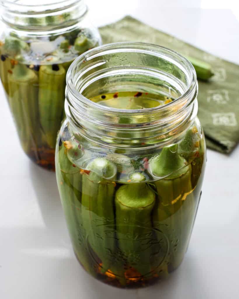 Two mason jars full of pickled okra with a green towel and fresh okra in the background.