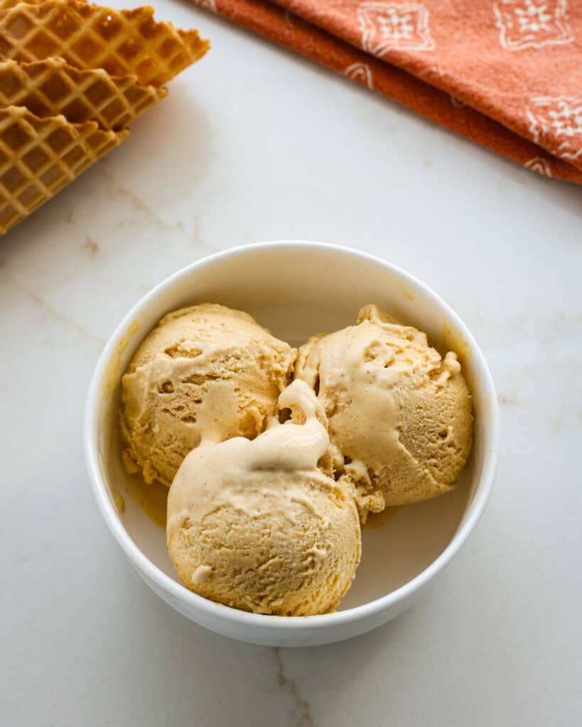 Three scoops of pumpkin ice cream in a white serving bowl with a few waffle cones in the background. 