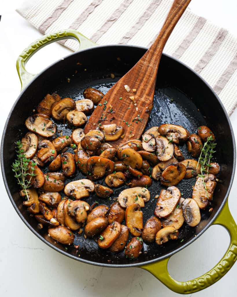 An overhead view of a cast iron skillet and wooden spatula full of sauteed mushrooms.