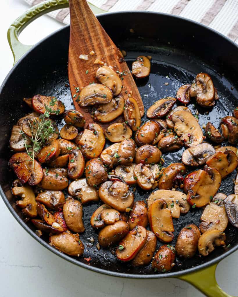 A close up view of a skillet and wooden spatula full of sauteed mushrooms.