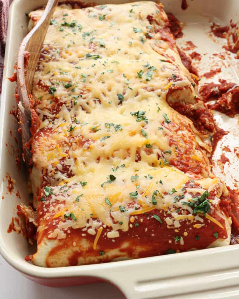 A close up view of wet burritos in a casserole dish with a wooden spatula.