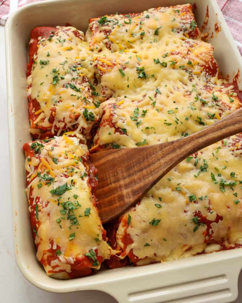 A casserole full of wet burritos with one of the burritos on a wooden spatula.