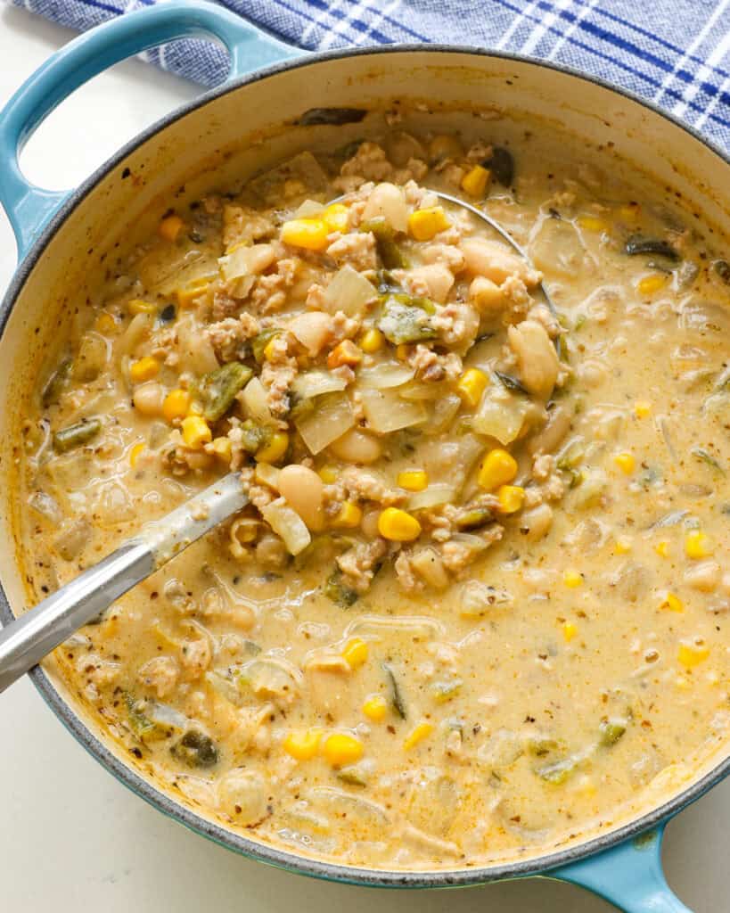 An overhead view of a Dutch oven and ladle full of white turkey chili with cannellini beans and roasted poblano peppers/ 